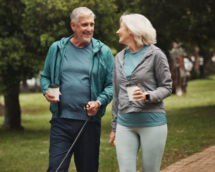 An older couple are walking their dog through a park trail while drinking coffee.